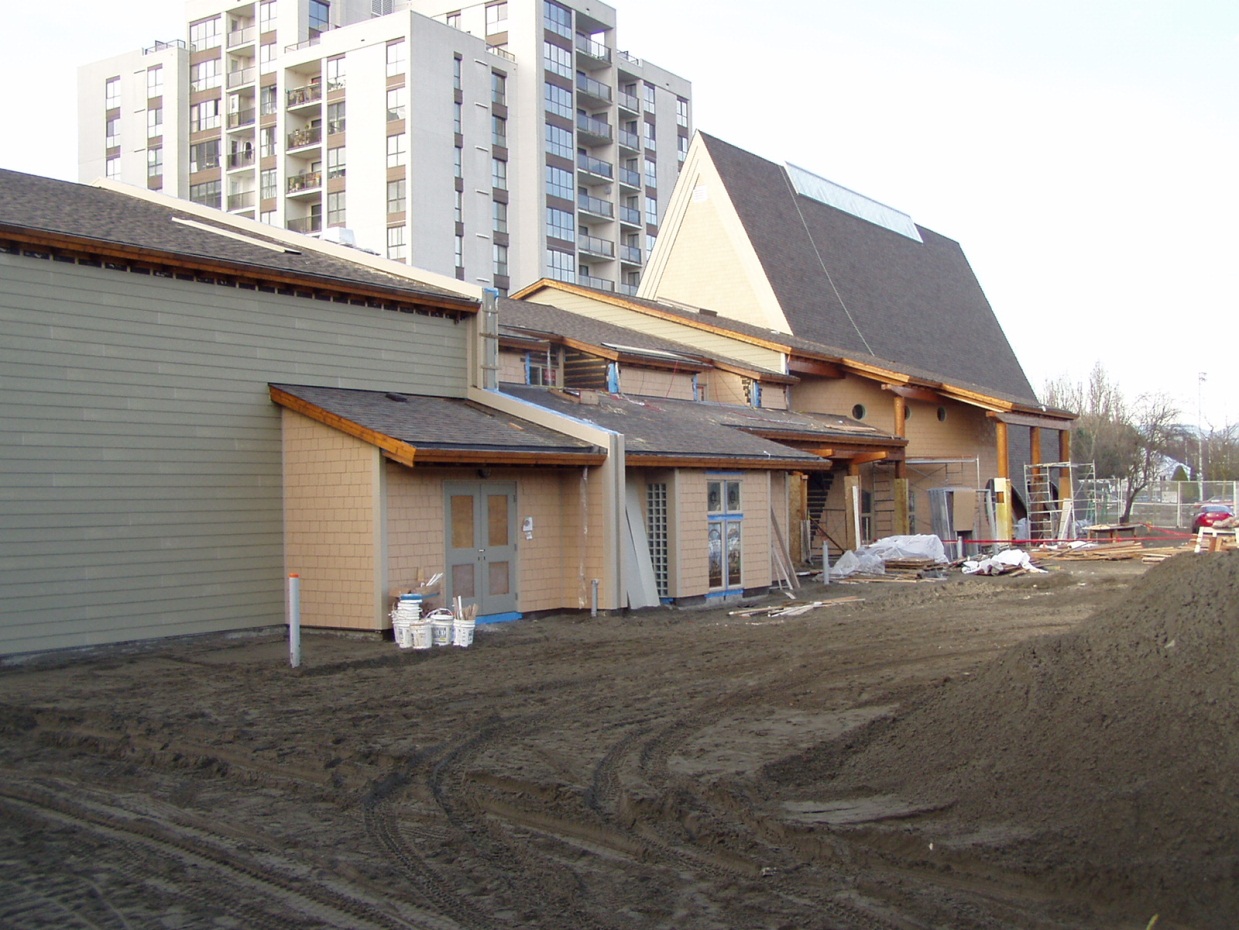 A 2003 photograph showing the detailed exterior finishes of the new multi-use facility at Trinity Lutheran Church in Richmond, BC. The image highlights the installation of horizontal lap siding and cedar-style shingles on the multi-use facility, with the 1964 A-frame sanctuary and its new skylight visible in the background against a newly graded soil surface.