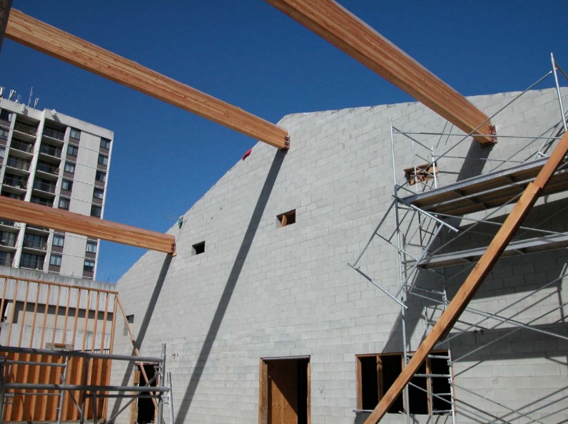 A photograph showing the concrete block masonry wall and large timber beams for the new parish hall at the south end of the multi-use facility at Trinity Lutheran Church in Richmond, BC. Scaffolding is positioned against the wall as the structural roof beams are set into place.