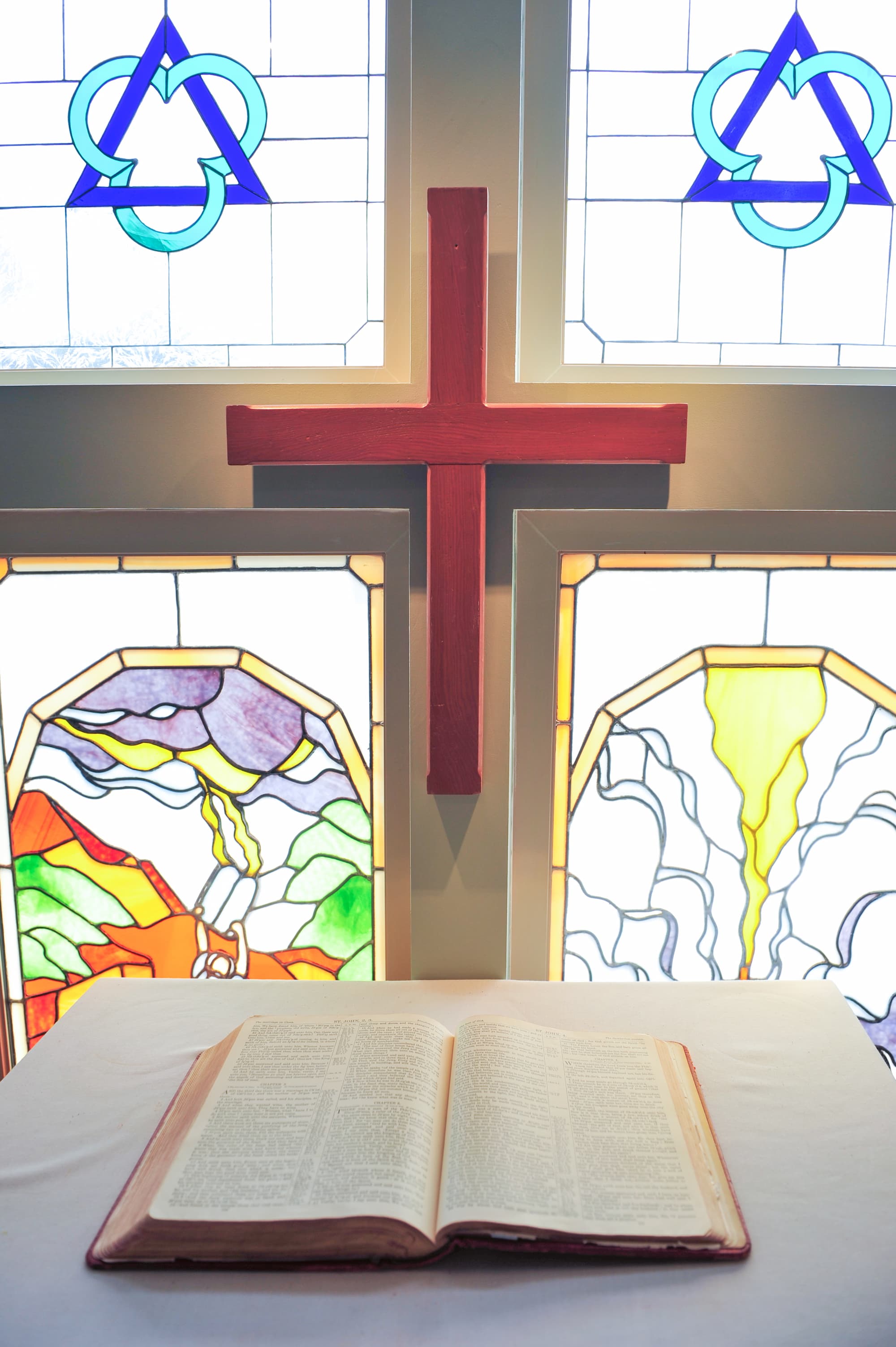 An open Holy Bible resting on a white altar cloth in the chapel. Behind the altar is a wooden cross and stained-glass panels depicting the Holy Trinity, Moses at Mt. Sinai (the Law), and the Crucifixion at Mt. Calvary (the Gospel).